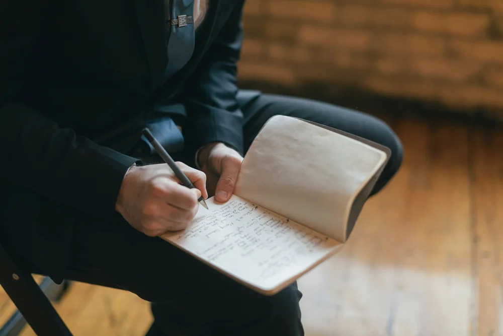 Personne assise de profil, sans visage visible, qui note à la main dans un carnet posé sur ses genoux
